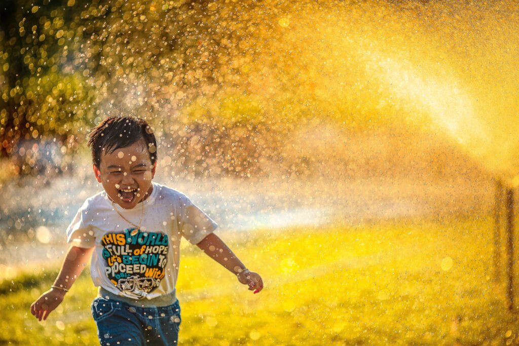 Young male toddler running through sprinkler in front yard at sunset.